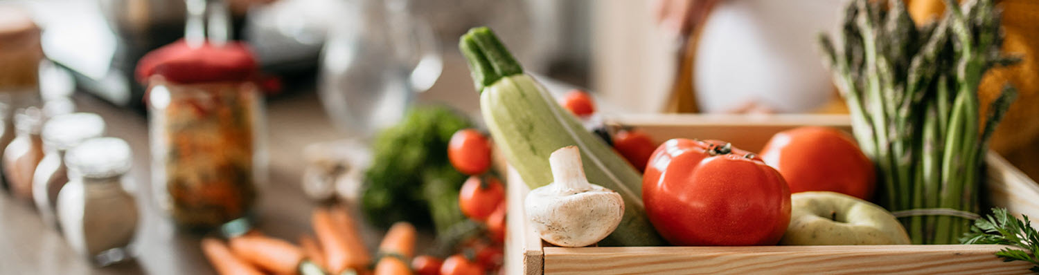 A wooden crate filled with fresh vegetables and a glass jar with a red lid.