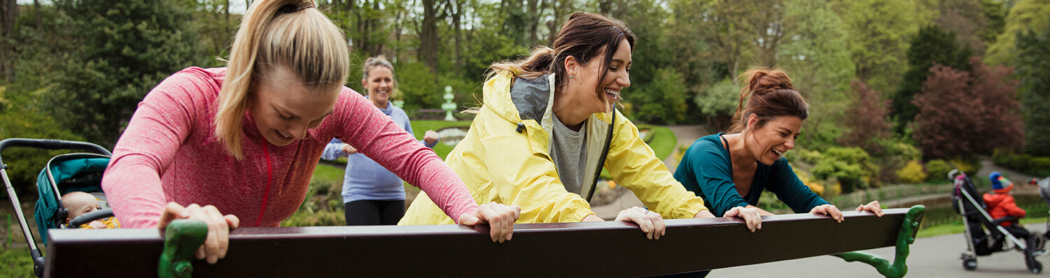 Four women are outdoors, leaning on a bench, with one woman pushing a stroller in the background.