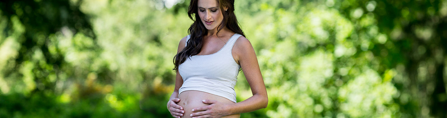 A pregnant woman in a white tank top stands outdoors, her hands gently resting on her belly.