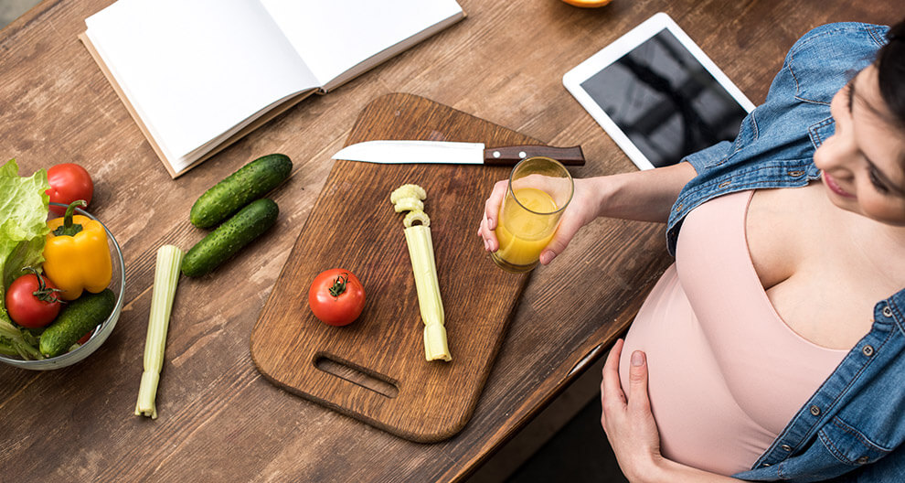 A pregnant woman is preparing a salad with fresh vegetables, holding a glass of orange juice, and using a tablet and book for reference.